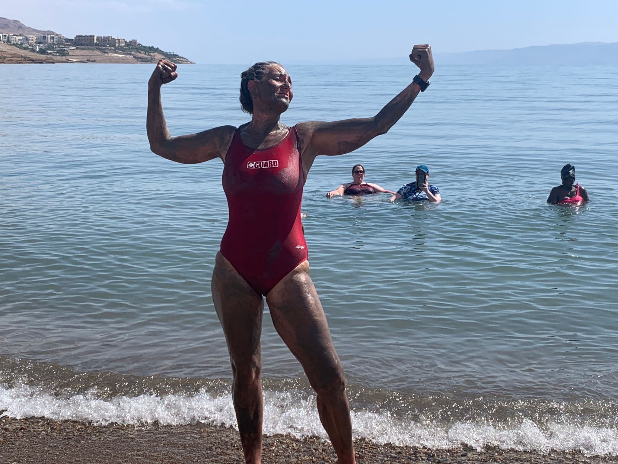 Woman in a red lifeguard swimsuit flexing on a pebbled shore with three people wading in calm water behind her