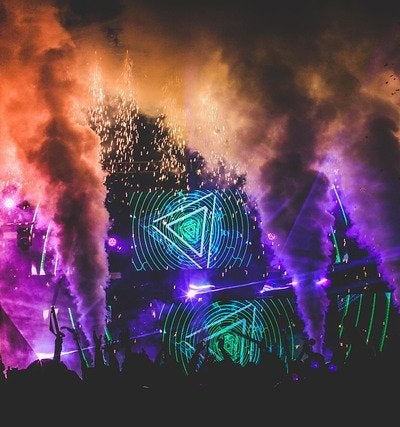 A massive music festival stage illuminated by vibrant purple and green laser beams cutting through atmospheric smoke