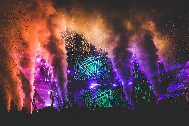 A massive music festival stage illuminated by vibrant purple and green laser beams cutting through atmospheric smoke