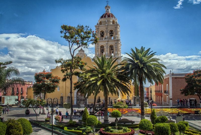 A vibrant Mexican plaza featuring a historic yellow church tower surrounded by lush palm trees and traditional colonial architecture