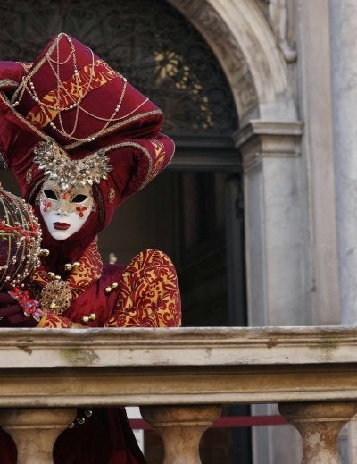 Person wearing ornate red and gold Venetian carnival mask with feathers standing on a balcony