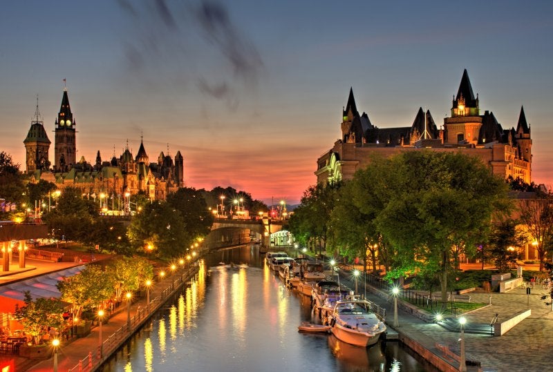 A beautiful warm orange-red glow fills the sky at dusk over Ottawa Canada during a July evening. A small wisp of low level clouds are visible. Parliament Hill, Chateau Laurier hotel and the Rideau Canal a UNSECO world heritage site are in the foreground.