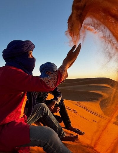 Two men in blue turbans sitting on a dune as one throws a large arc of sand on the Sahara Desert at sunset.
