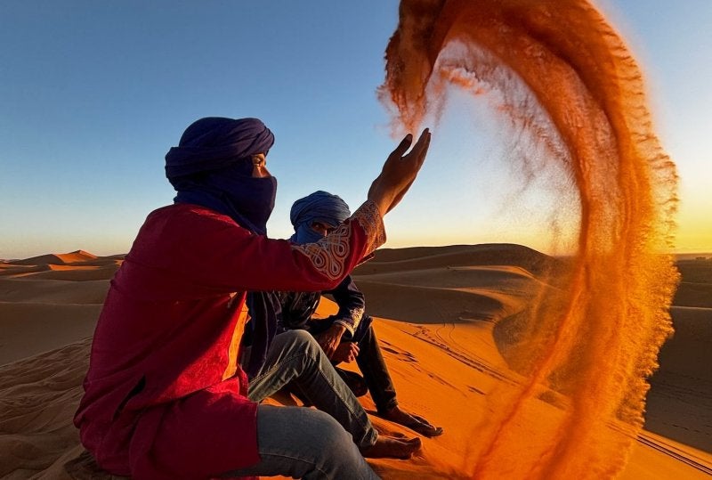 Two men in blue turbans sitting on a dune as one throws a large arc of sand on the Sahara Desert at sunset.