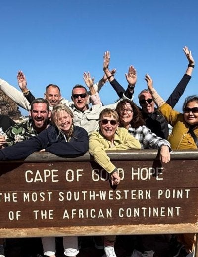 Cape of Good Hope sign with a group of travelers leaning on it and cheering by the rocky shore in South Africa.
