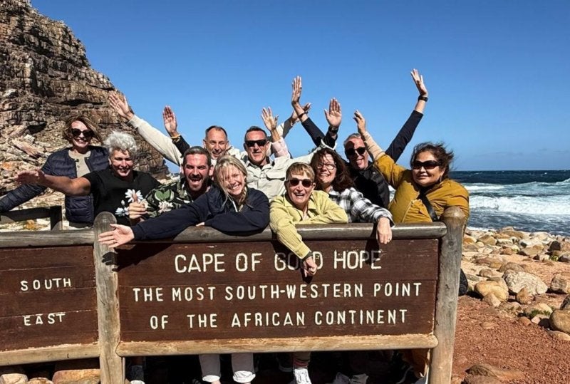Cape of Good Hope sign with a group of travelers leaning on it and cheering by the rocky shore in South Africa.